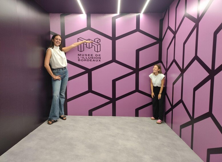 Two people in a room with a purple geometric wall design and a logo for 'MUSEE DE L'ILLUSION BORDEAUX.' 3