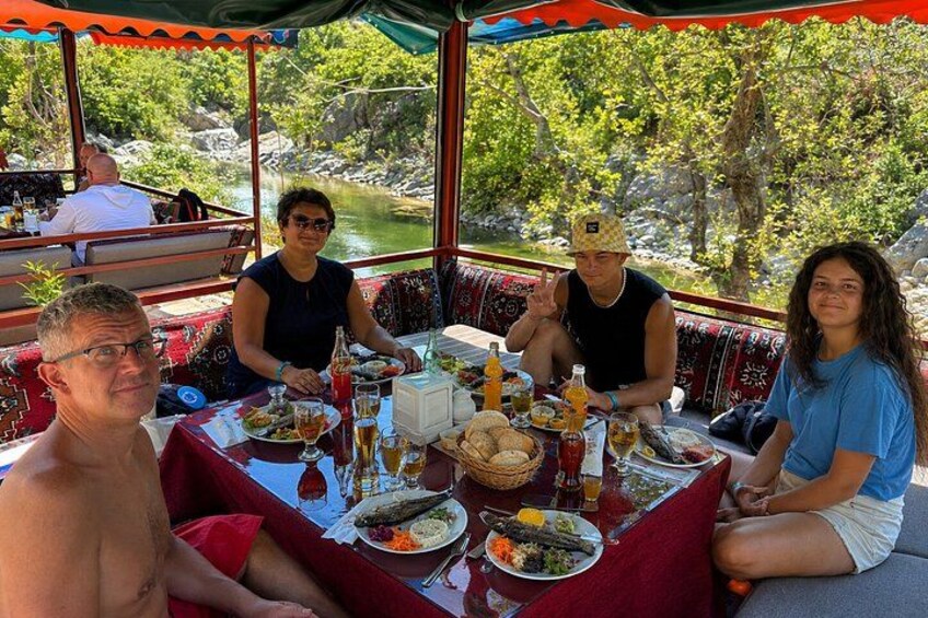 A group of people dining at an outdoor table with a river and greenery in the background. 5