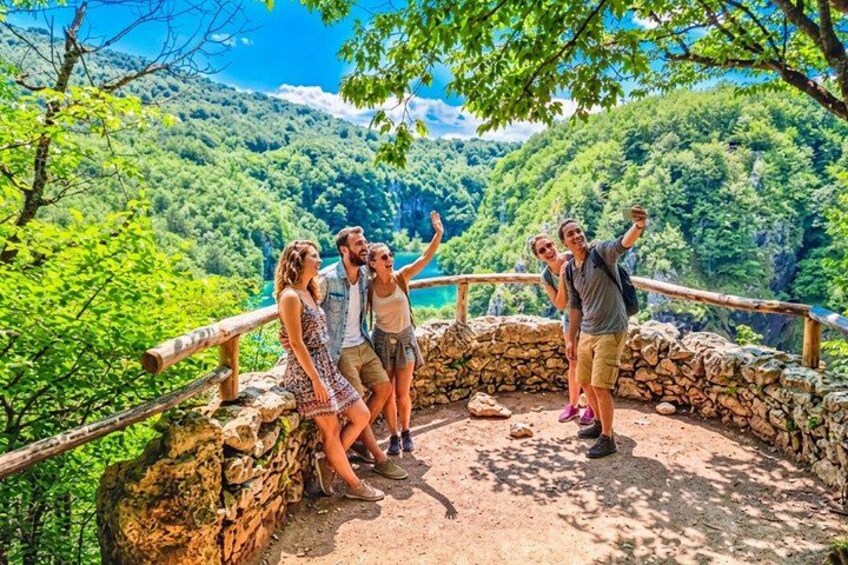 A group of people standing on a stone platform with a wooden railing, overlooking a lush green valley and a body of water. 5