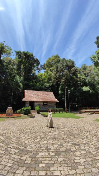 A woman standing in a courtyard with a stone floor, surrounded by trees and a building with a red roof. 4