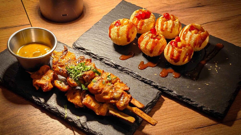 Skewers of grilled meat, a bowl of sauce, and a slate plate with small pastries and sauce. 2