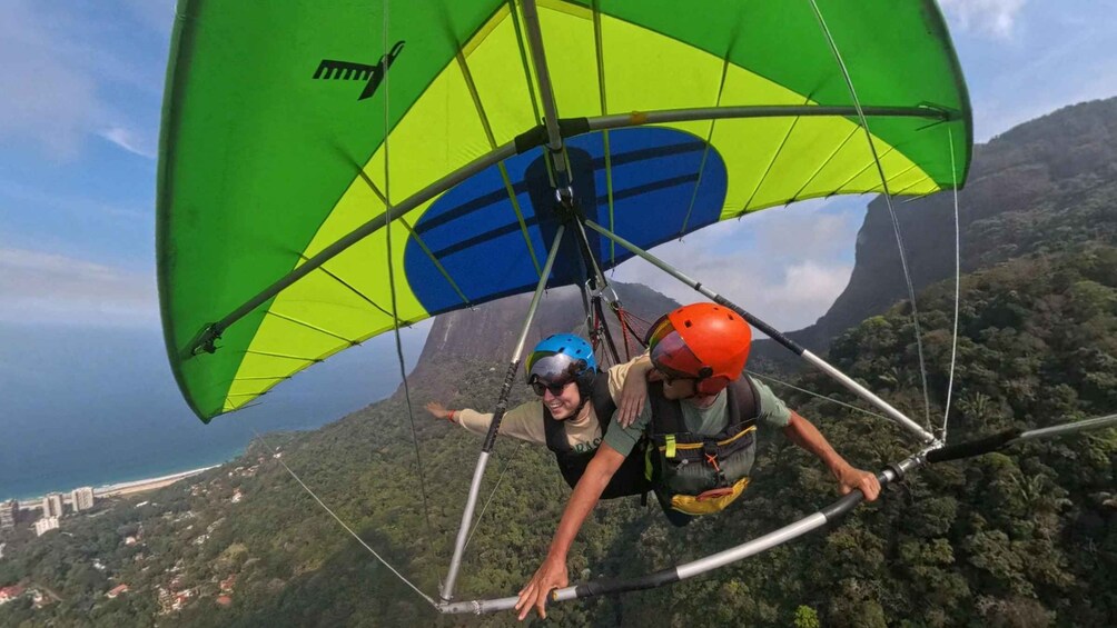 Two people paragliding with a green and blue canopy, over a mountainous landscape with a view of the ocean and buildings in the distance. 4