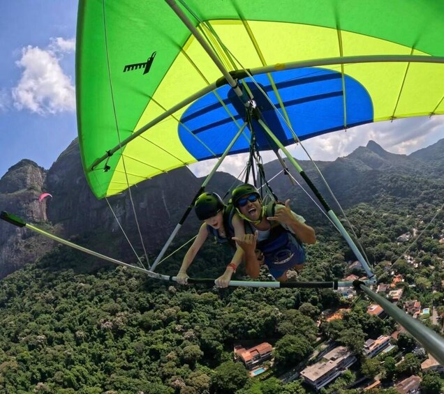 A person in a hang glider with a green and blue canopy flying over a forested area with mountains in the background. 4