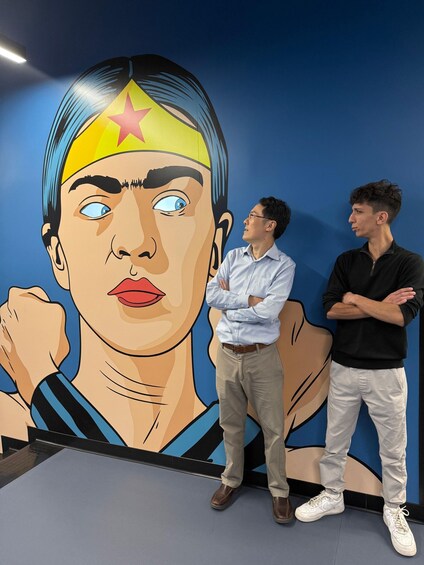 A mural of a female superhero with a star on her head, two men standing in front of it, one with arms crossed. 1
