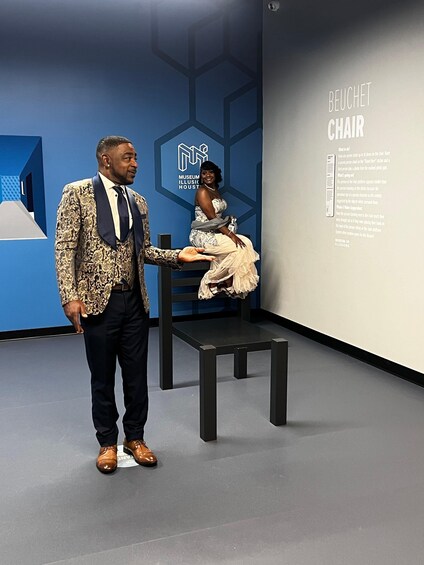 A man and a woman in a museum exhibit with a chair labeled 'BEUCHET CHAIR' and a hexagonal wall design. 5