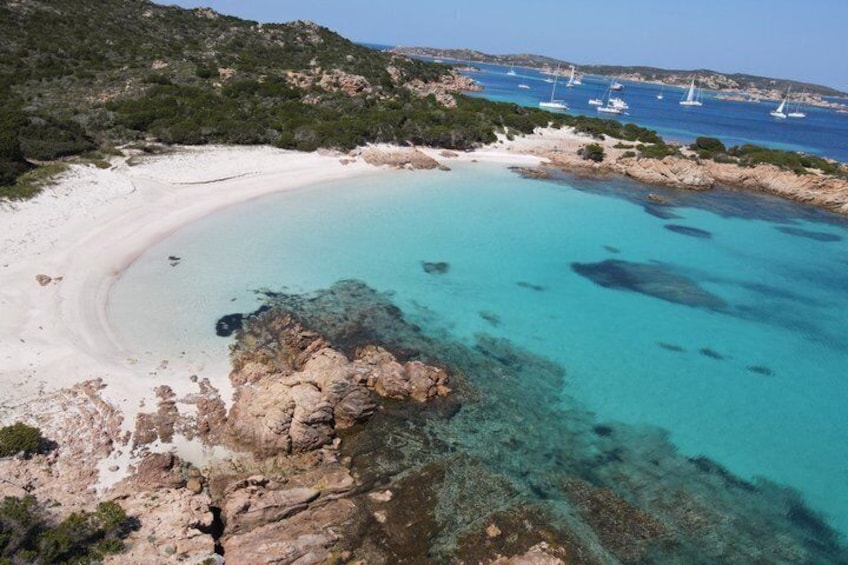 A coastal landscape with a sandy beach, clear turquoise waters, rocky outcrops, and sailboats in the distance. 4