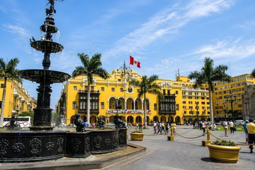 A fountain, palm trees, and a yellow building with a flag. 1