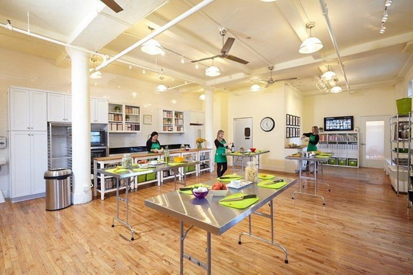A spacious kitchen with wooden floors, white cabinets, and stainless steel appliances. There are three people working at a long table with food items and cooking utensils. 2