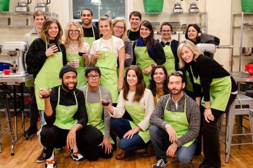 A group of people in a kitchen wearing aprons, standing and sitting around kitchen equipment. 1