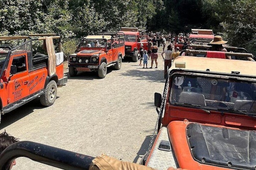 A line of orange safari vehicles parked on a gravel surface with people standing in the background. 5