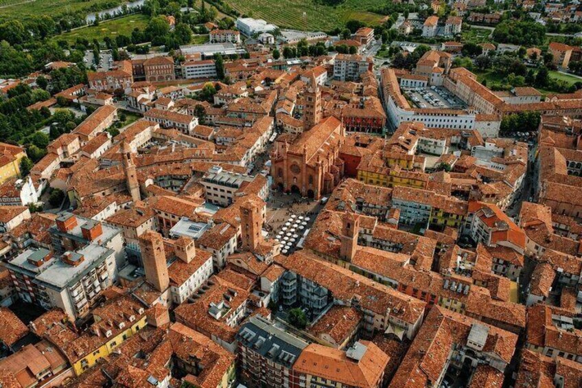 An aerial view of a historic town with dense architecture, prominent towers, and surrounding greenery. 1