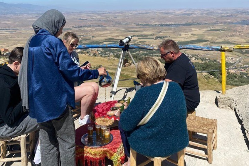 A group of people sitting at a table with a telescope, overlooking a valley. 4