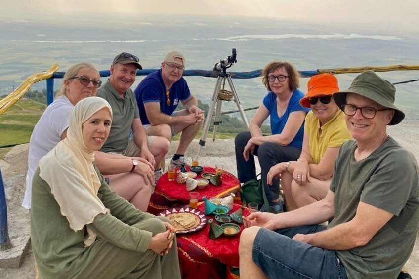 A group of people sitting around a table with a telescope on a high vantage point overlooking a landscape. 3