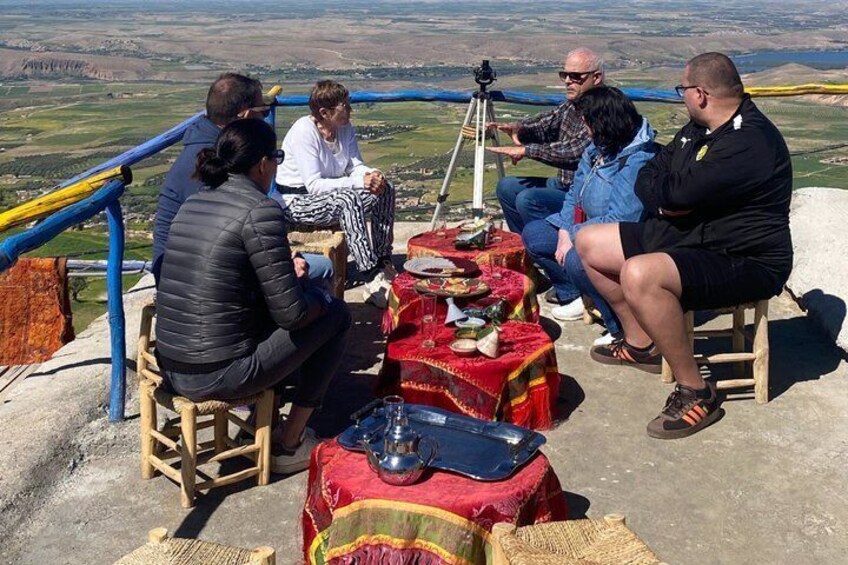 A group of people sitting on stools at a table with a camera on a tripod, overlooking a valley. 2