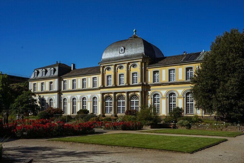 A large, historic building with a prominent dome and symmetrical architecture, surrounded by well-maintained gardens and a clear blue sky. 1