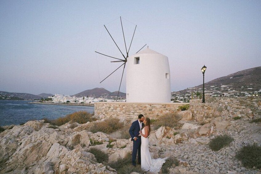 A couple embracing near a white windmill by the sea, with rocky terrain and a distant town. 2