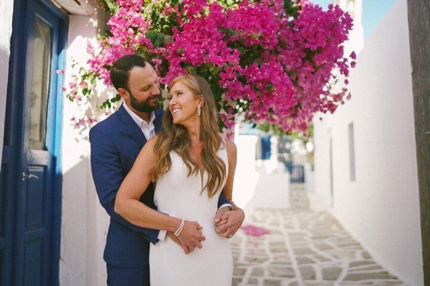 A couple standing in a narrow street with white buildings and pink bougainvillea flowers. 3