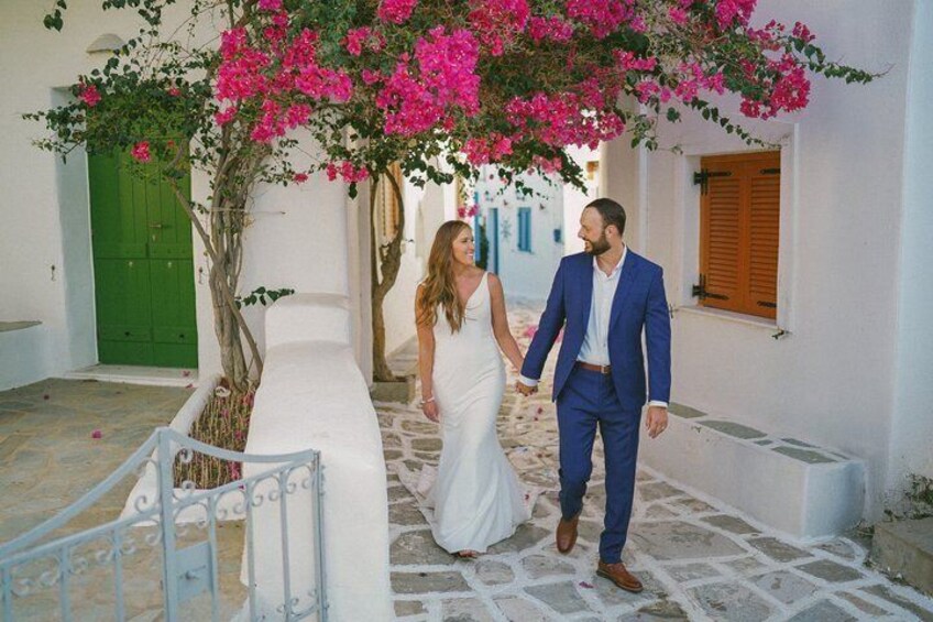 A couple walking on a white-washed street with pink bougainvillea flowers, a green door, and a white building. 4