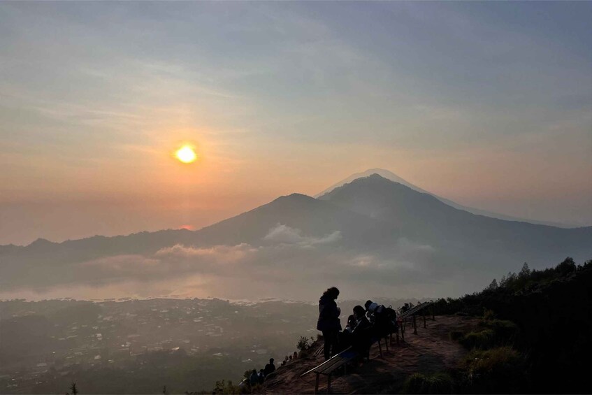A mountain with a sunrise, a person standing, and a group of people on a viewing platform. 5