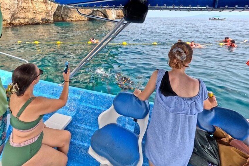 Two people on a boat, one taking a photo, with clear water and rocky cliffs in the background. 5