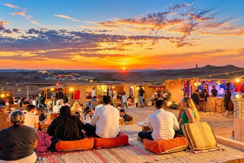 A sunset over a desert landscape with people sitting on cushions. 2