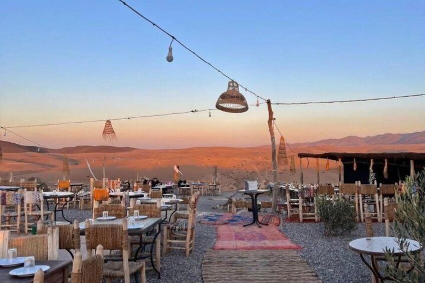 An outdoor dining area with tables and chairs, a patterned rug, and hanging lights against a backdrop of a desert landscape. 2