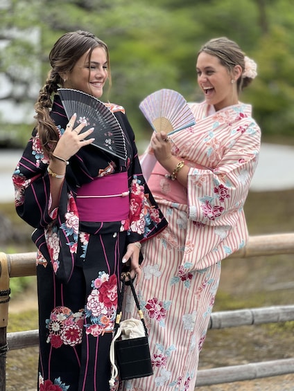 Two individuals in traditional Japanese attire, one holding a fan and the other a decorative fan, standing by a wooden fence. 2