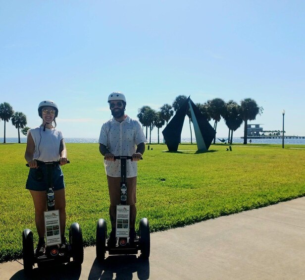 Two people on electric scooters in a park with a sculpture and palm trees. 3