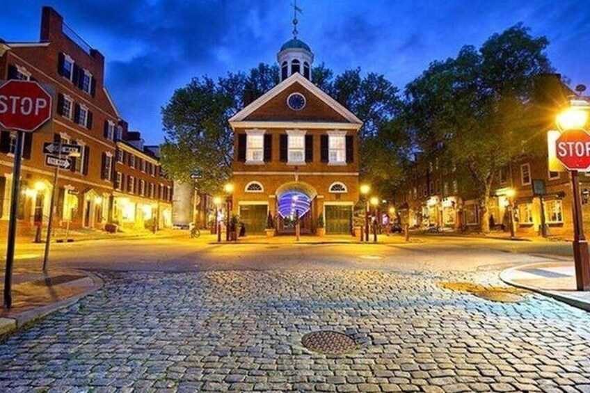 A cobblestone street leading to a historic building with a clock tower, surrounded by brick buildings and illuminated street lamps. 1