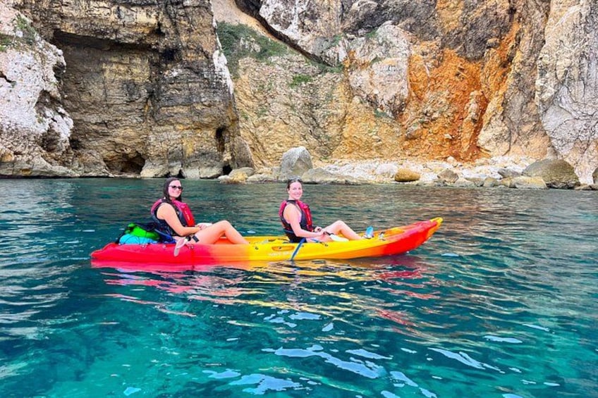Two people kayaking on a clear blue sea with rocky cliffs in the background. 2