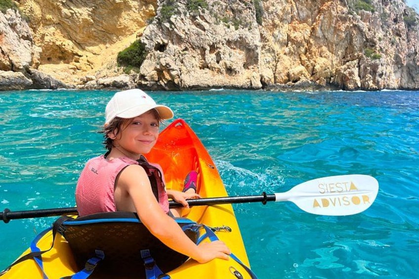 A person in a yellow kayak with a life jacket, holding a paddle, on clear blue water with rocky cliffs in the background. 3