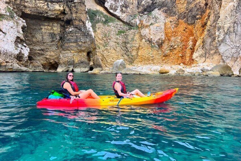 Two people kayaking on a clear blue water body with rocky cliffs in the background. 2