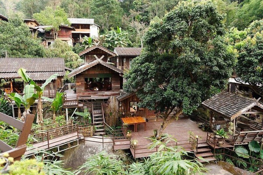 A wooden house with a staircase, surrounded by lush greenery and other wooden structures. 2