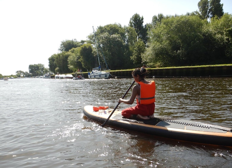 A person on a paddleboard in a calm river with boats and greenery in the background. 1