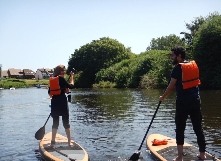 Two people paddleboarding on a calm river, wearing life jackets, surrounded by lush greenery and houses in the background. 2