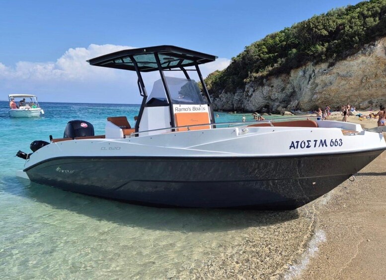 A speedboat named 'Ramo's Boats' with the registration 'ATOE TM 663' docked at a beach with clear water and rocky cliffs in the background. 1
