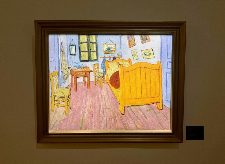 A framed painting of a room with a bed, a table, chairs, and pictures on the wall. 2