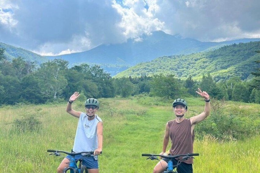 Two people on bicycles in a grassy field with mountains in the background. 3