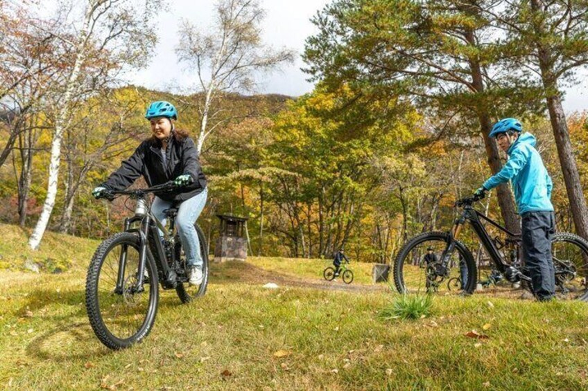 Two cyclists on a trail in a wooded area with autumn foliage. 1