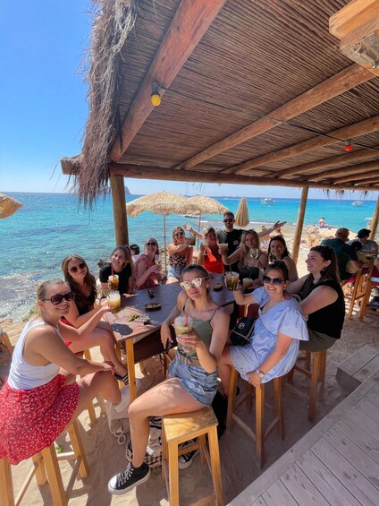 A group of people sitting at wooden tables under a thatched roof, overlooking a beach with the sea and boats in the background. 2