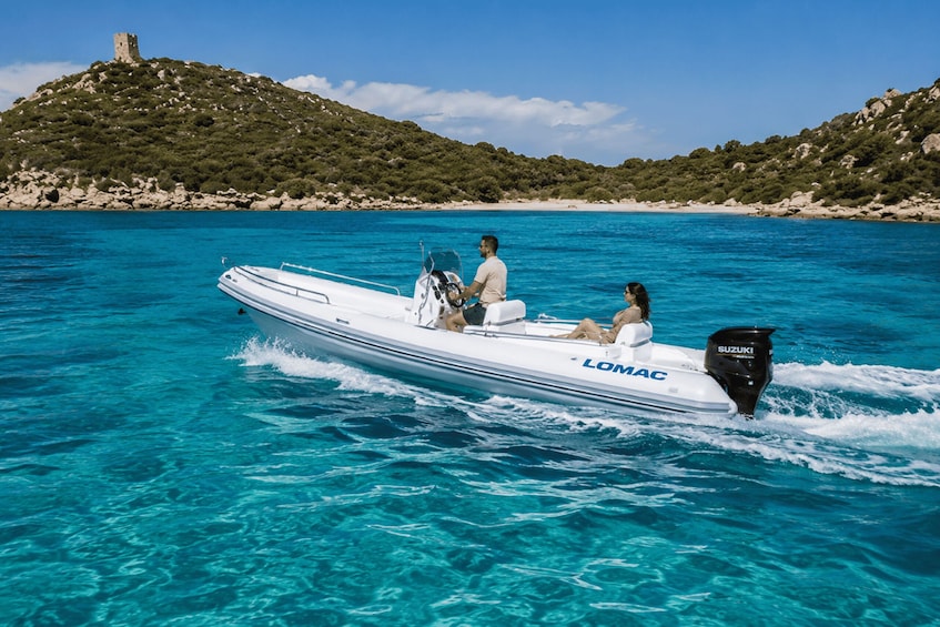 A boat named 'LOMAC' on clear turquoise water with a rocky hill and a tower in the background. 1