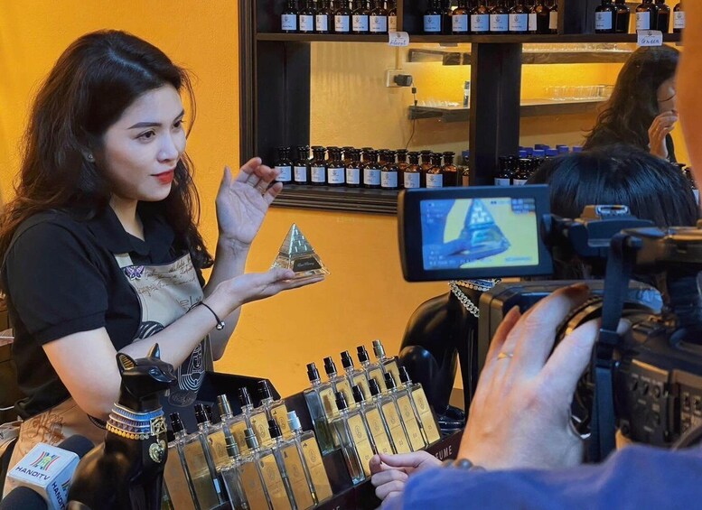 A person holding a perfume bottle in a store with shelves of other bottles and a camera in the foreground. 1