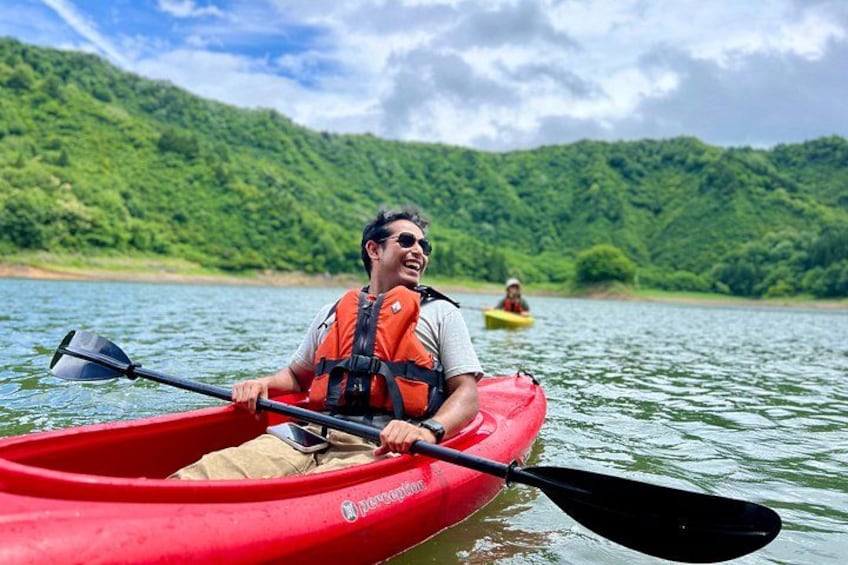A person kayaking on a river with a red kayak, another person in a yellow kayak in the background, and green hills in the distance. 1