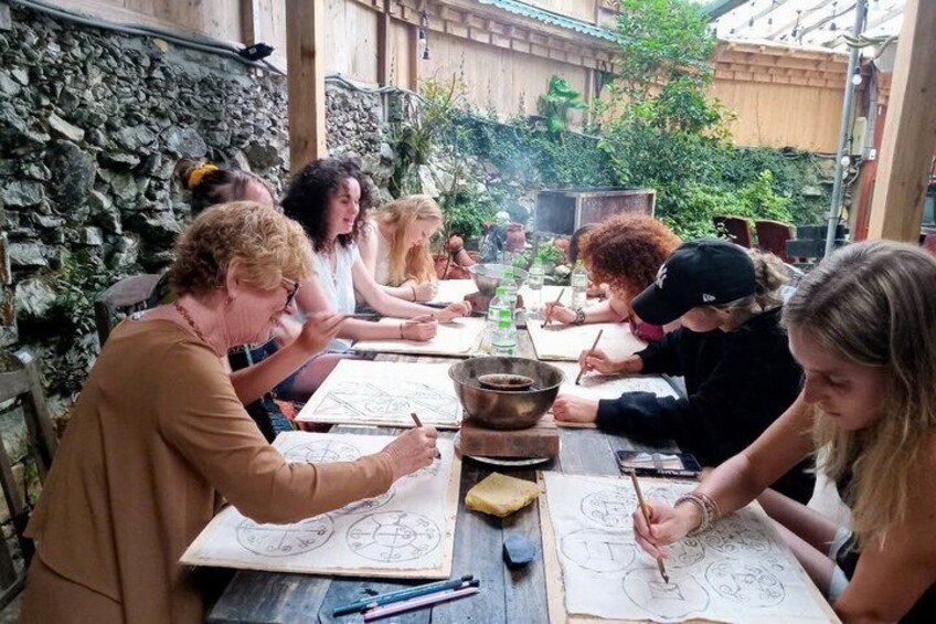A group of people engaged in a drawing activity outdoors, seated around a table with drawing materials. 3