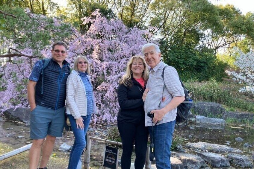 Four people standing in front of cherry blossoms with a pond and rocks in the background. 2