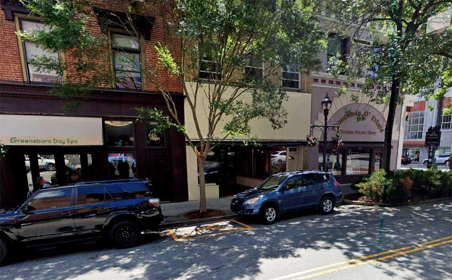 A street with parked cars, a brick building, and a sign for 'Greenboro Day Spa.' 2