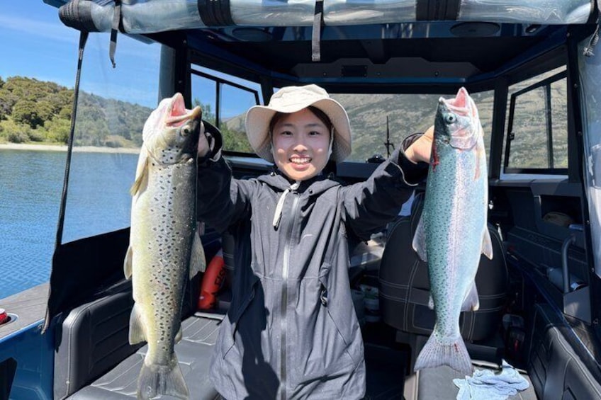 A person holding two fish on a boat with a lake and hills in the background. 4