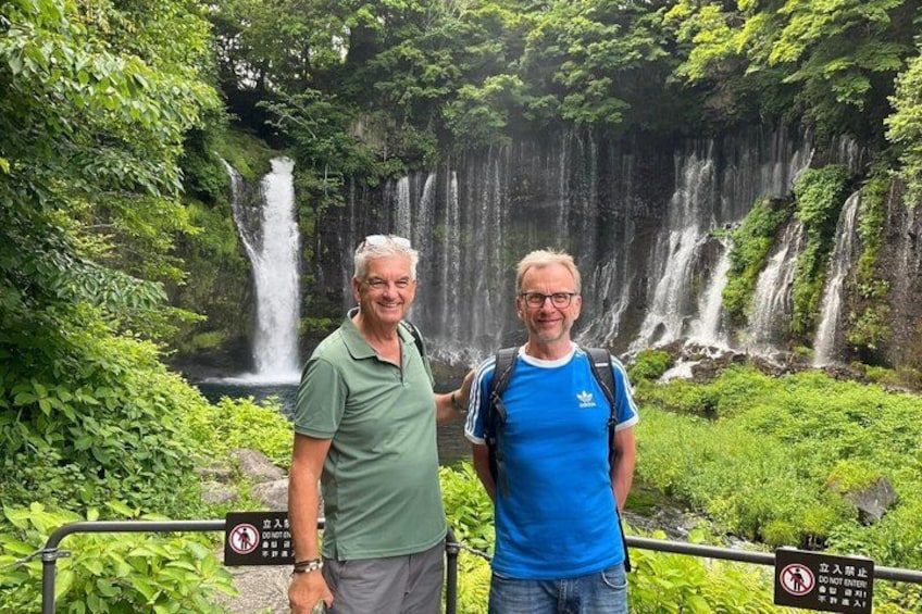 Zwei Personen stehen vor einem Wasserfall, umgeben von üppigem Grün. 4