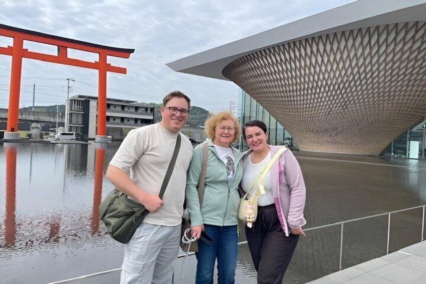 Three people standing in front of a modern building with a distinctive roof and a red torii gate in the background. 5