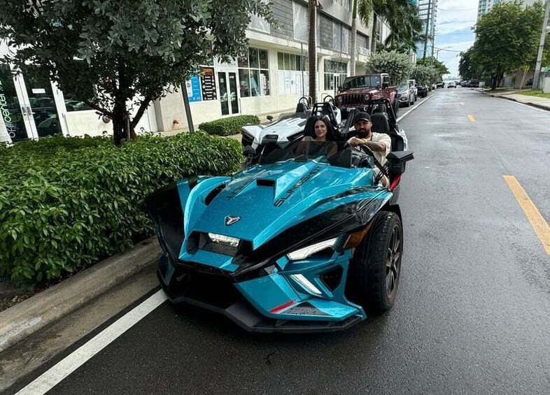 A blue sports car parked on a city street with buildings and trees in the background. 5